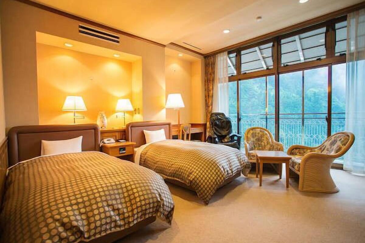A hotel room with two beds, a small table, two chairs, a lamp, and a view of the outdoors.