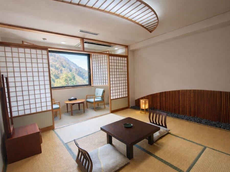 Una habitación japonesa tradicional con suelo de tatami, una mesa baja, sillas, una puerta corredera y vistas a la montaña.