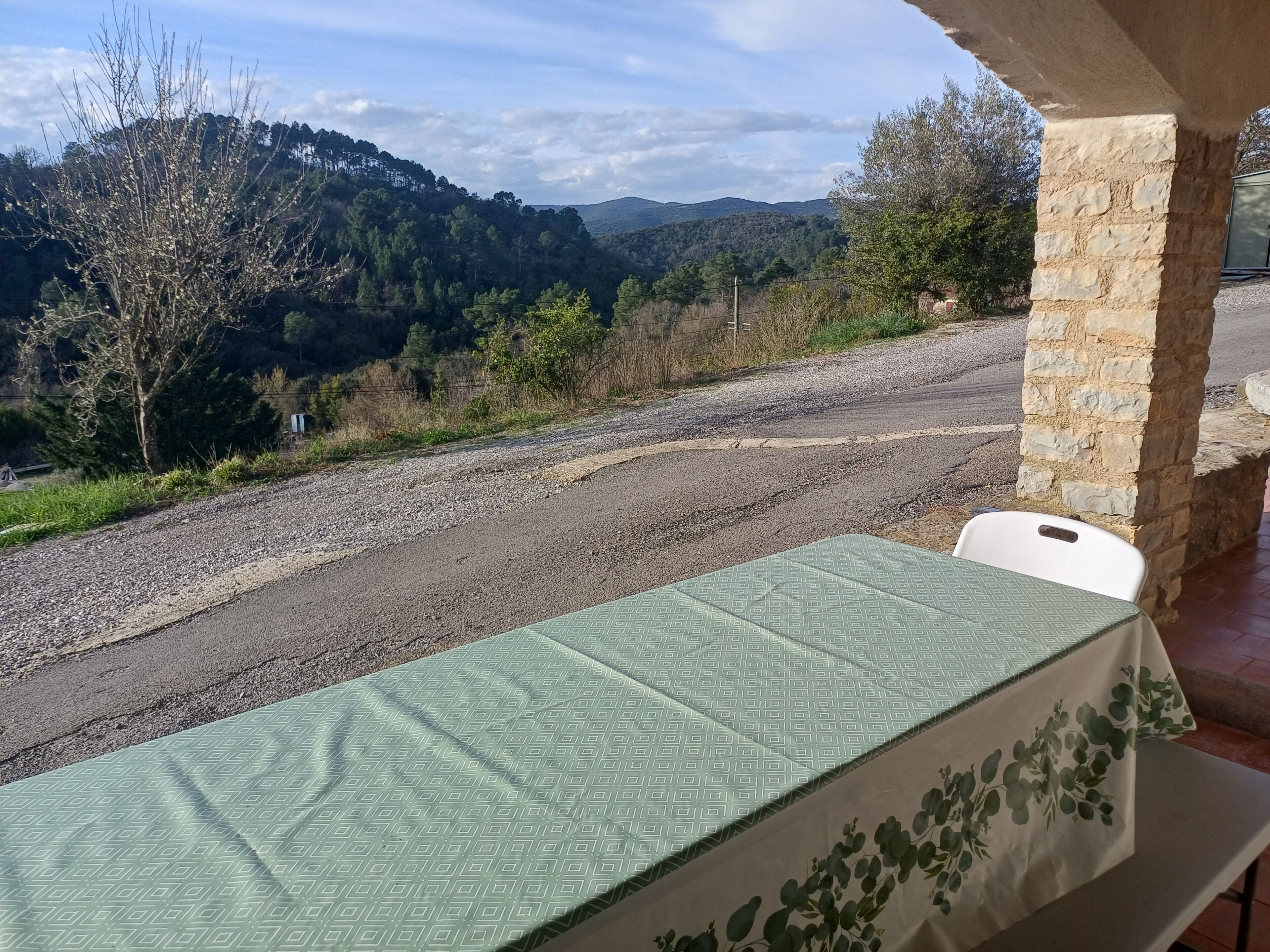 Une table recouverte d’une nappe à motifs, un pilier en pierre et une vue sur un chemin de gravier menant à une zone boisée.