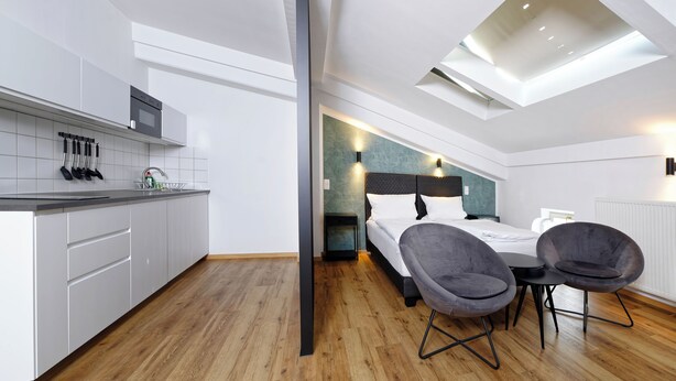 A modern hotel room with a kitchen, a bed with a headboard, two chairs, and a skylight. - Boardinghouse Rosenheim (Rosenheim)