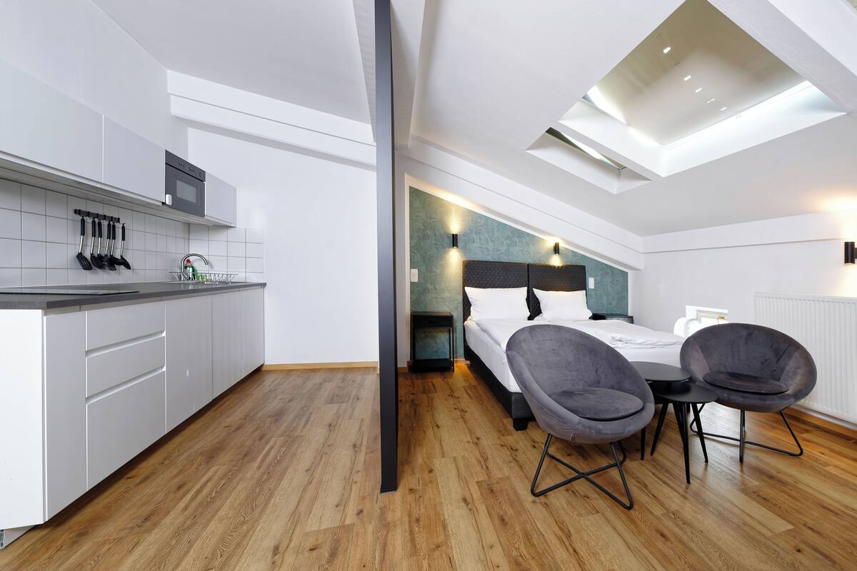A modern hotel room with a kitchen, a bed with a headboard, two chairs, and a skylight.