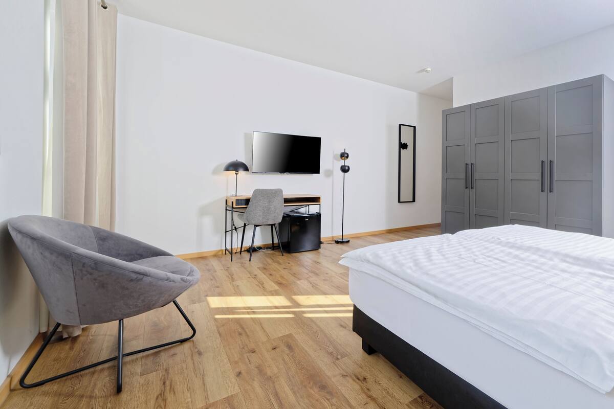 A modern hotel room with a bed, a desk with a chair, a television, and a wardrobe.