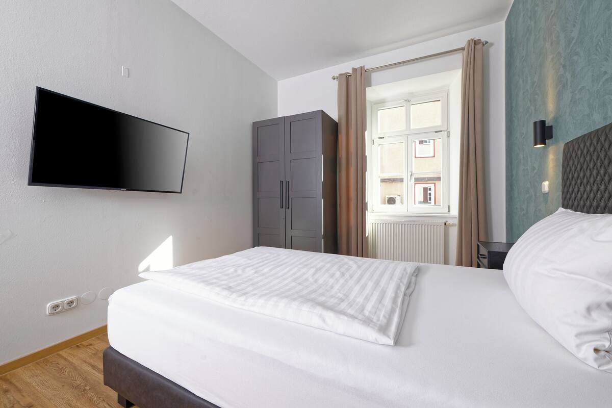 A hotel room with a bed, a flat-screen TV mounted on the wall, a wardrobe, and a window with curtains.