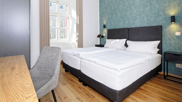 A hotel room with a bed, a chair, a nightstand, a lamp, and a window with curtains. - Boardinghouse Rosenheim (Rosenheim)