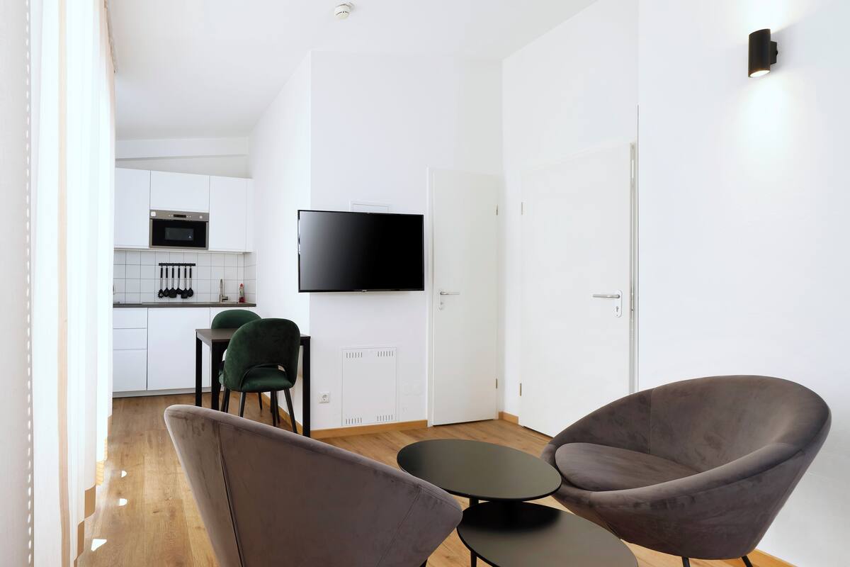 A modern living room with a dining area, a flat-screen TV, and a kitchen in the background.