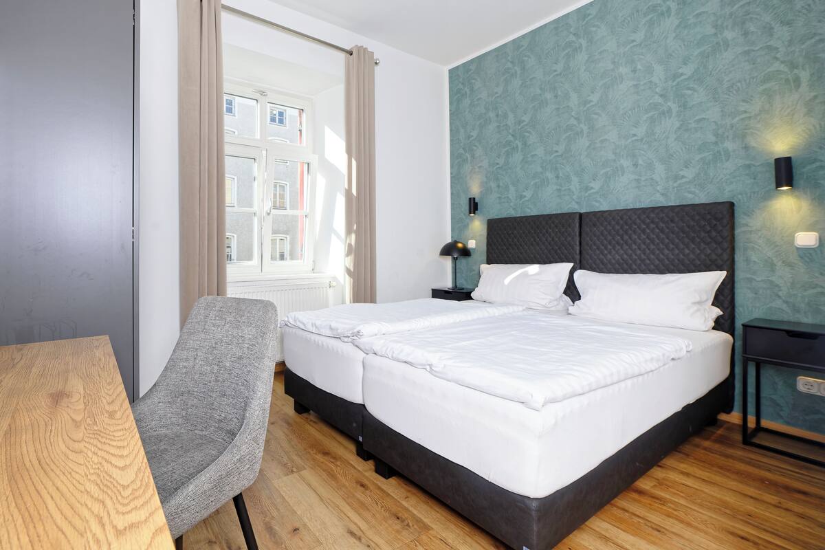 A hotel room with a bed, a chair, a nightstand, a lamp, and a window with curtains.