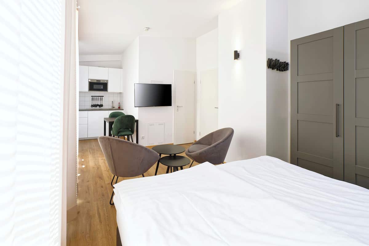 A hotel room with a bed, two armchairs, a small table, a TV, a kitchen area, and a wardrobe.