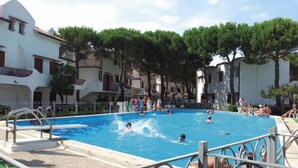 A swimming pool surrounded by buildings with balconies, trees, and a clear sky. - Flat in seafront residence with pool (Rosolina Mare)