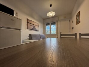 Ein Zimmer mit Holzboden, einer Couch, einem Tisch, einem Kühlschrank, einem Fenster mit Jalousien und einer Deckenleuchte.