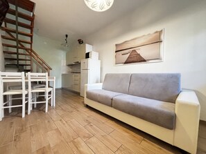 Ein Wohnzimmer mit einer Couch, einem Esstisch mit Stühlen, einer Treppe, einer Küchenzeile und einem an der Wand hängenden Bild.