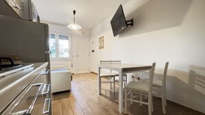 Cocina compacta con refrigerador, una mesa de comedor con sillas, televisor montado en la pared y una puerta que da a otra habitación.