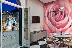A dining area with a view of a cityscape through a window, a round table with chairs, a vase with flowers, and a wall with a large rose mural. - Lady Rose One (Naples)