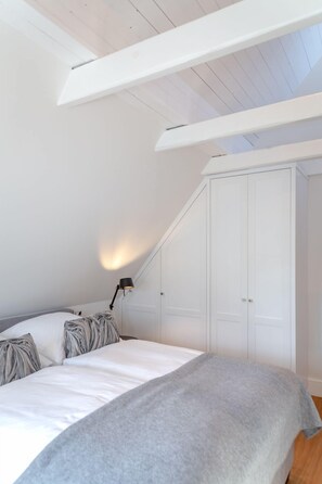Une chambre avec un plafond en pente, un lit avec des oreillers à rayures blanches et grises, un plaid gris, une lampe de sol noire et une armoire blanche.