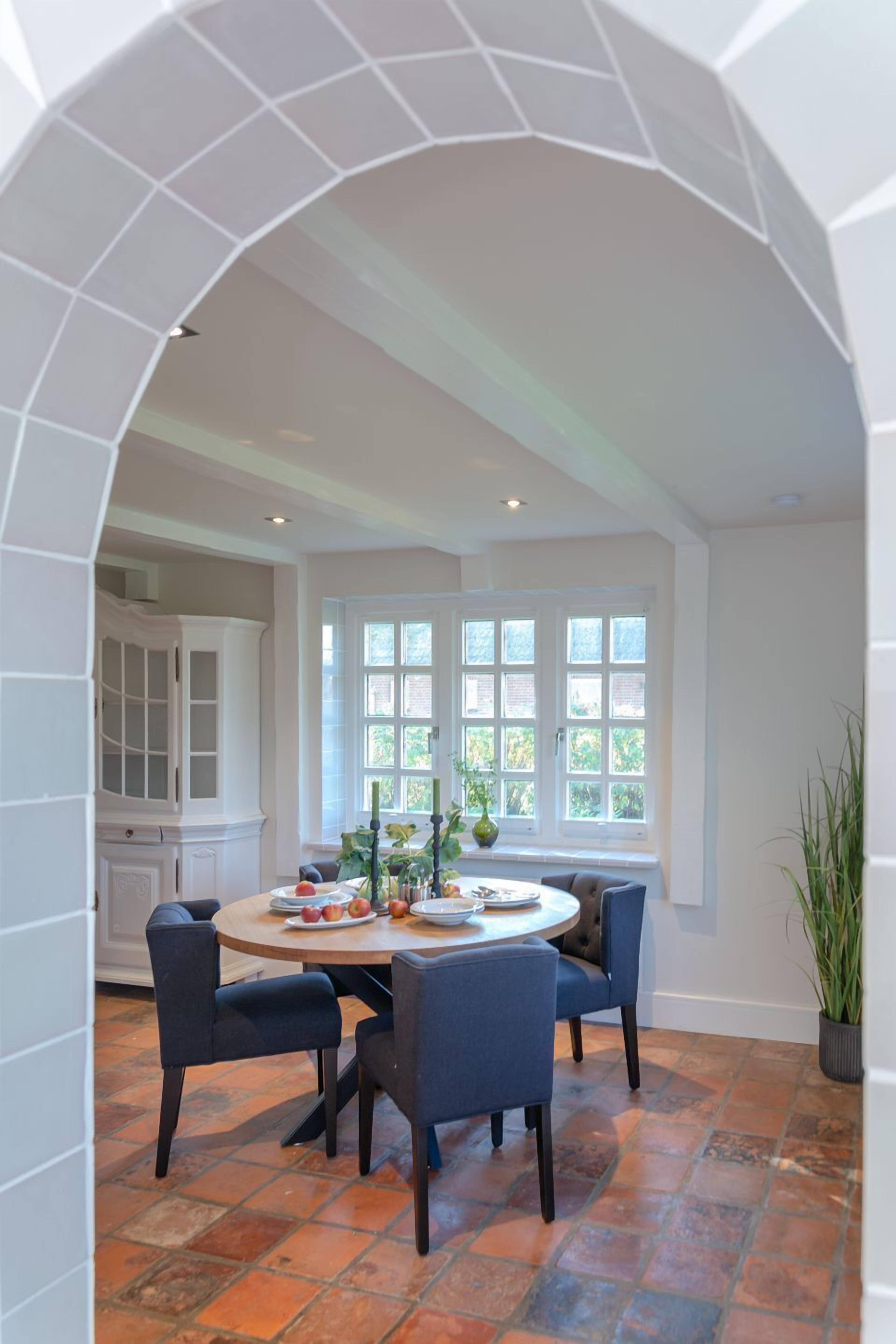 A dining area with a round table, four chairs, a window with multiple panes, and a white cabinet in the background.