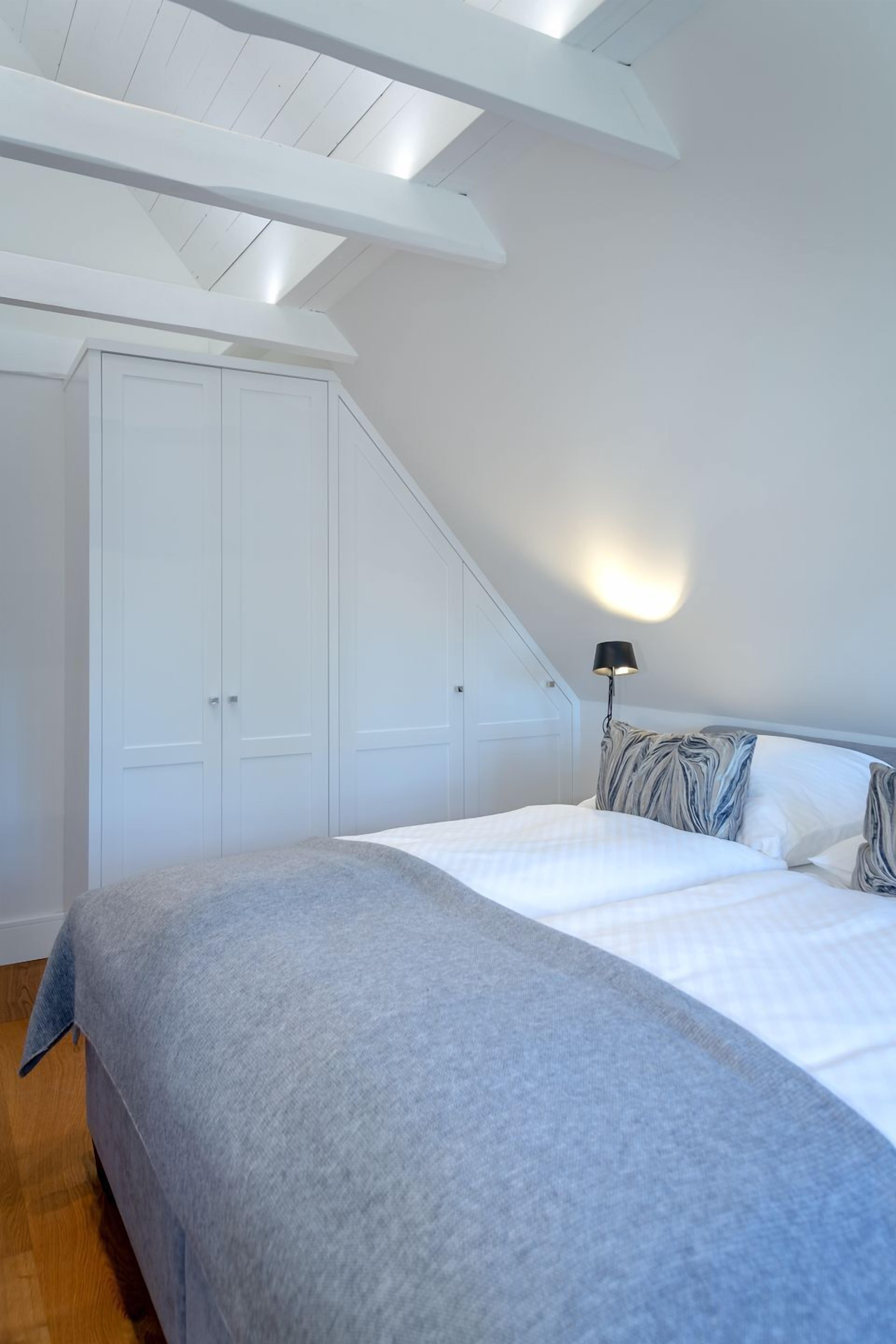 A bedroom with a sloped ceiling, a bed with a grey blanket, white pillows, and a black lamp.