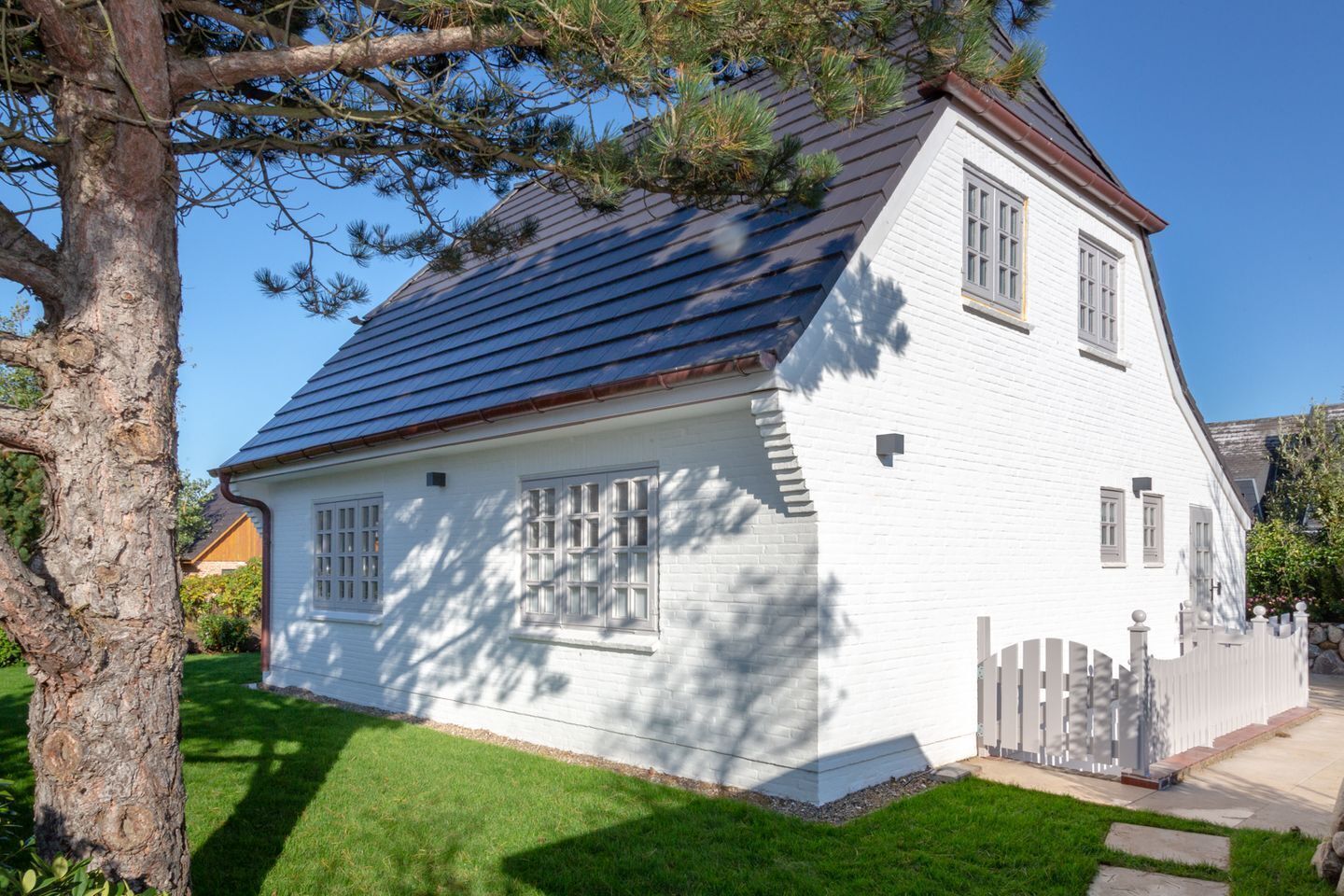 A white two-story house with a dark roof, a wooden fence, and a tree in the foreground.