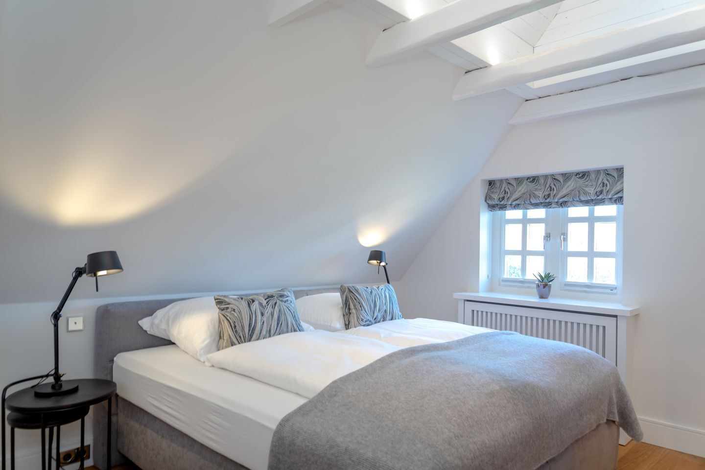 Ein Schlafzimmer mit einem Bett, zwei Nachttischlampen, einem Fenster mit Pflanze und einer Heizung.