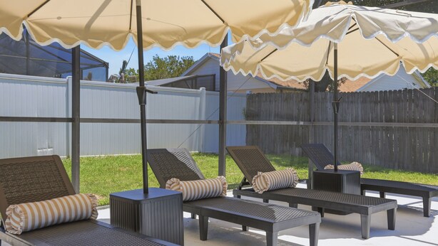 Une terrasse extérieure dotée de plusieurs chaises longues et de parasols, d’une table en métal et offrant une vue sur une cour arrière délimitée par une clôture en bois.