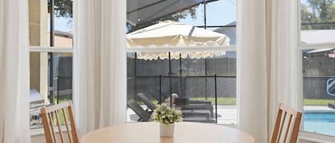 A round wooden table with chairs, a potted plant, and a view of a backyard with a pool and umbrellas.