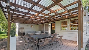 Un porche cubierto con una mesa y sillas de madera, una zona de estar con cojines y vistas a la casa.