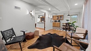 Amplia sala de estar con piso de madera, un sofá de cuero negro, un sillón de cuero marrón y un tapete de cuero negro. Cuenta con una zona de cocina con gabinetes de madera y piso a cuadros.