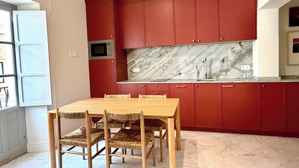 A kitchen with red cabinets, a marble backsplash, and a wooden dining table with woven chairs. - Alameda Grand Luxury (Seville)