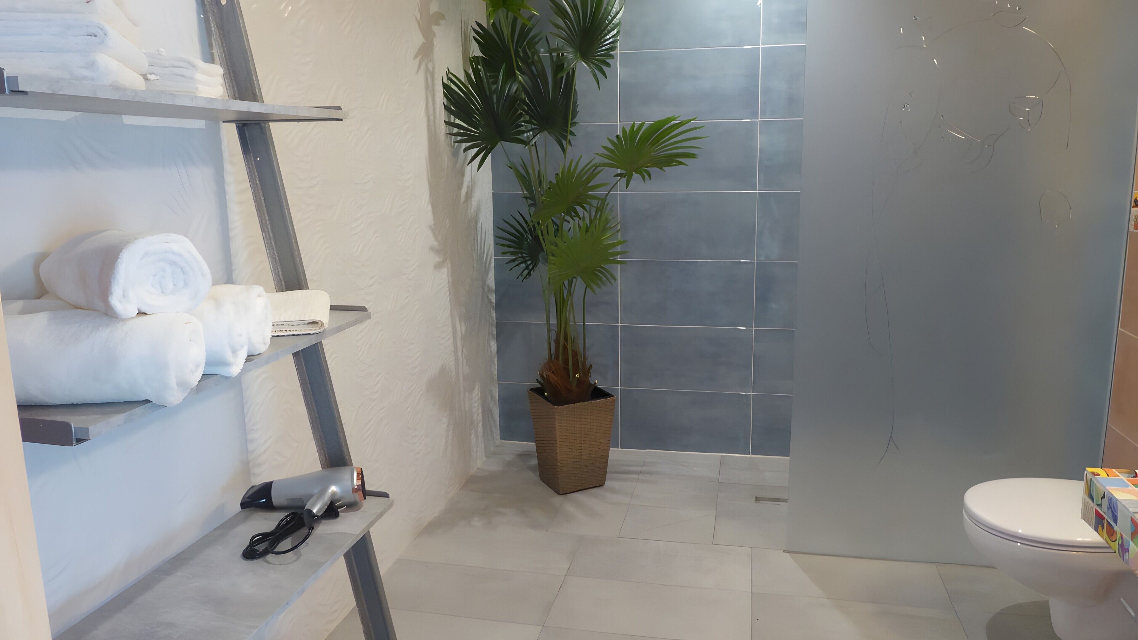 Un baño con ducha, inodoro, un estante con toallas, secador de pelo y una planta en maceta.