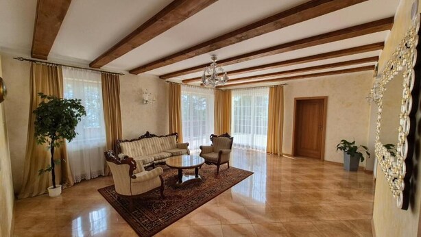 Amplia sala de estar con sofá, mesa de centro y sillas, vigas de madera en el techo, ventanas grandes con cortinas y una araña.