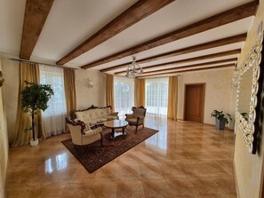 Uma sala de estar ampla com sofá, mesa de centro e poltronas, vigas de madeira no teto, janelas grandes com cortinas e um lustre.