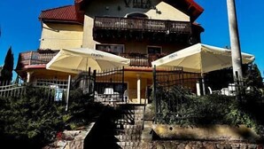 Un edificio con un letrero que dice 'Villa Mura', un balcón, asientos exteriores con sombrillas y una escalera de piedra que conduce a la entrada.