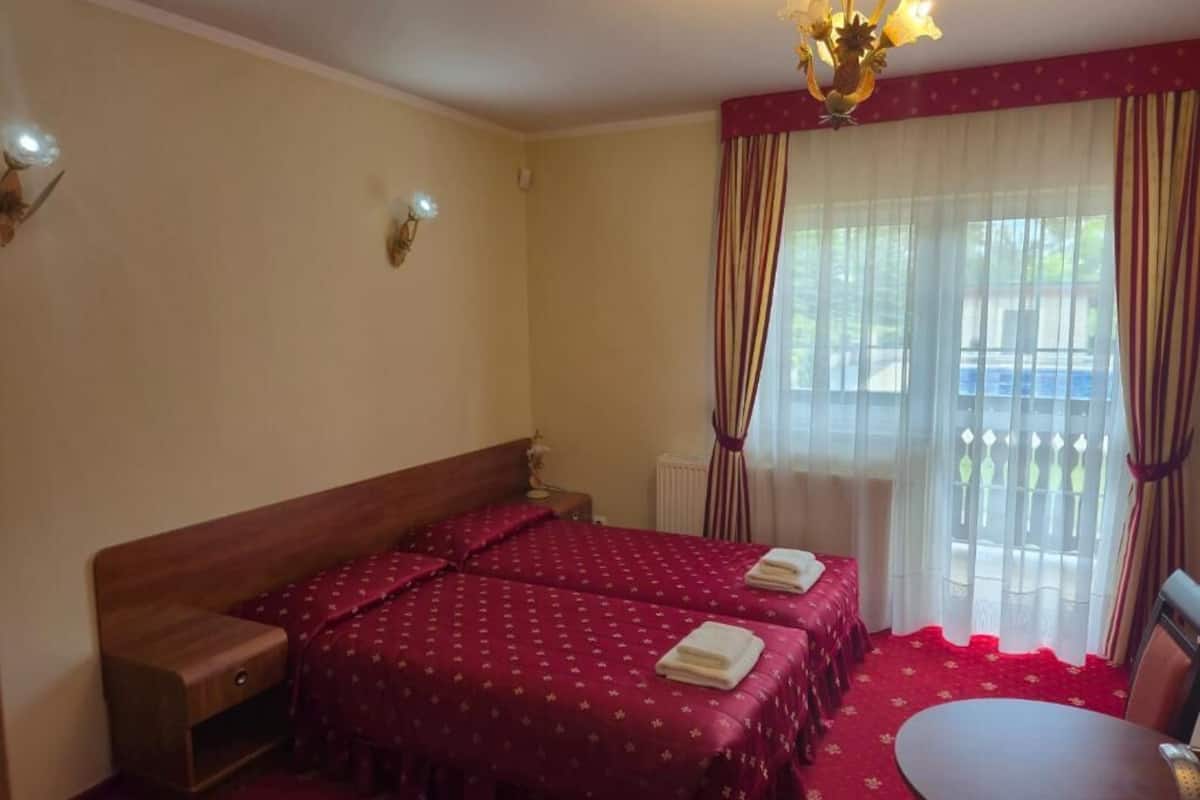 Habitación de hotel con dos camas, una mesita, una silla, un radiador y una ventana con cortinas.