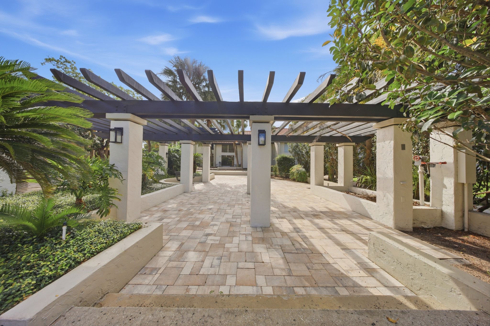 A covered walkway with a pergola structure, surrounded by landscaped gardens and palm trees.
