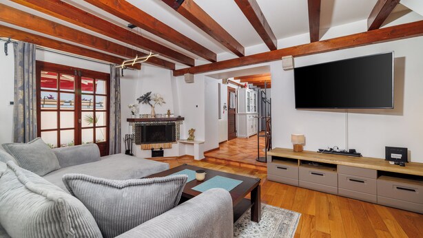 Ein Wohnzimmer mit einer Couch, einem Couchtisch, einem Flachbildfernseher, Holzbalken, einem Kamin und einem Flur, der zur Küche führt.