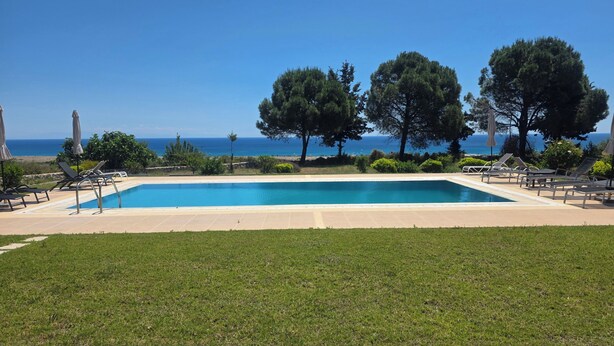En pool med liggestole og træer, med udsigt over et klart, blåt hav.