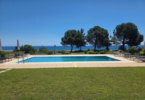 Piscina com espreguiçadeiras e árvores, com vista para um mar azul e límpido.