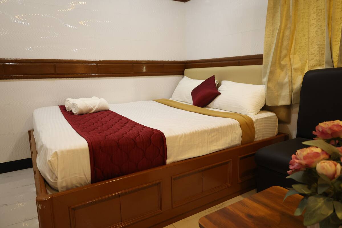 A hotel room with a wooden bed, a red blanket, a white pillow, and a vase with flowers on a wooden table.