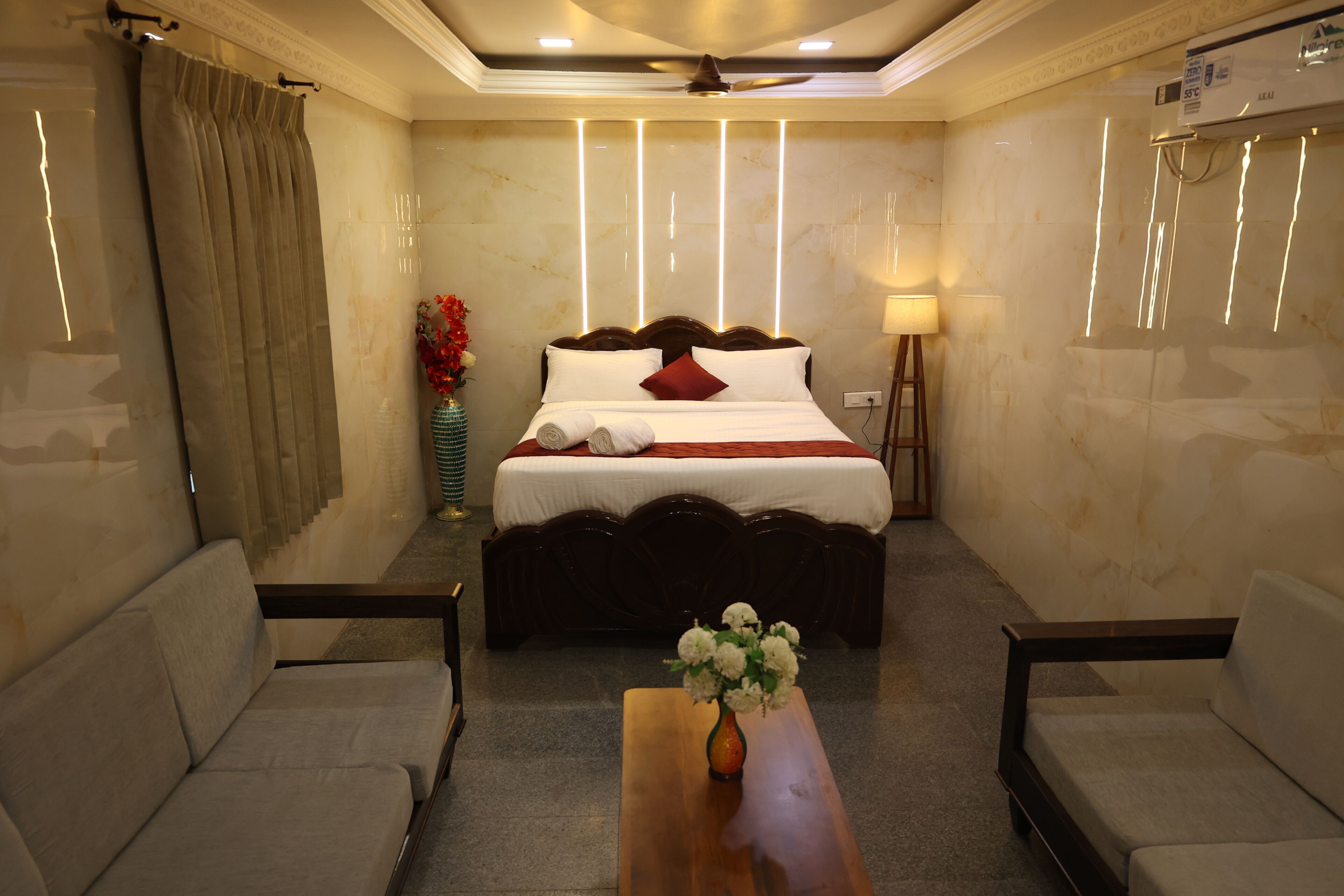 Habitación de hotel con una cama, dos sillones, una mesa de madera, un florero con flores y una lámpara de pie.