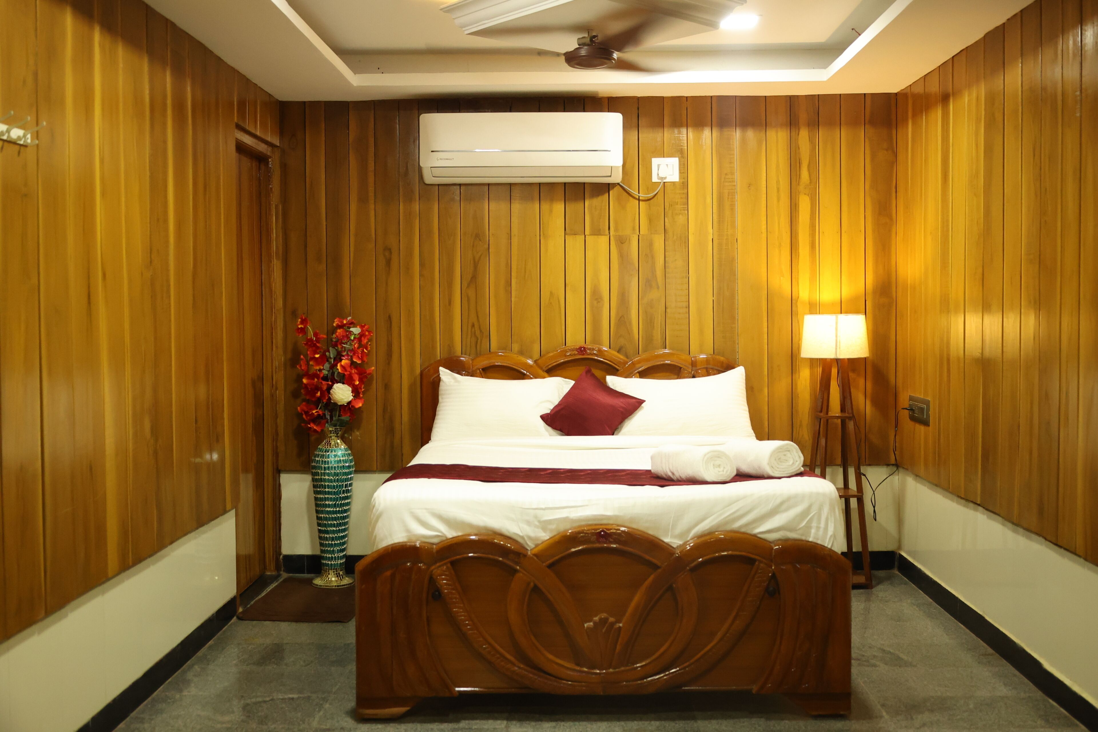 A hotel room with wooden panel walls, a bed with white linens, a vase with red flowers, and a standing lamp.