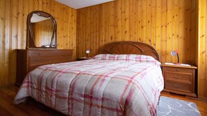 A bedroom with a wooden paneled wall, a bed with a plaid bedspread, a dresser with a mirror, and a bedside table with a lamp.