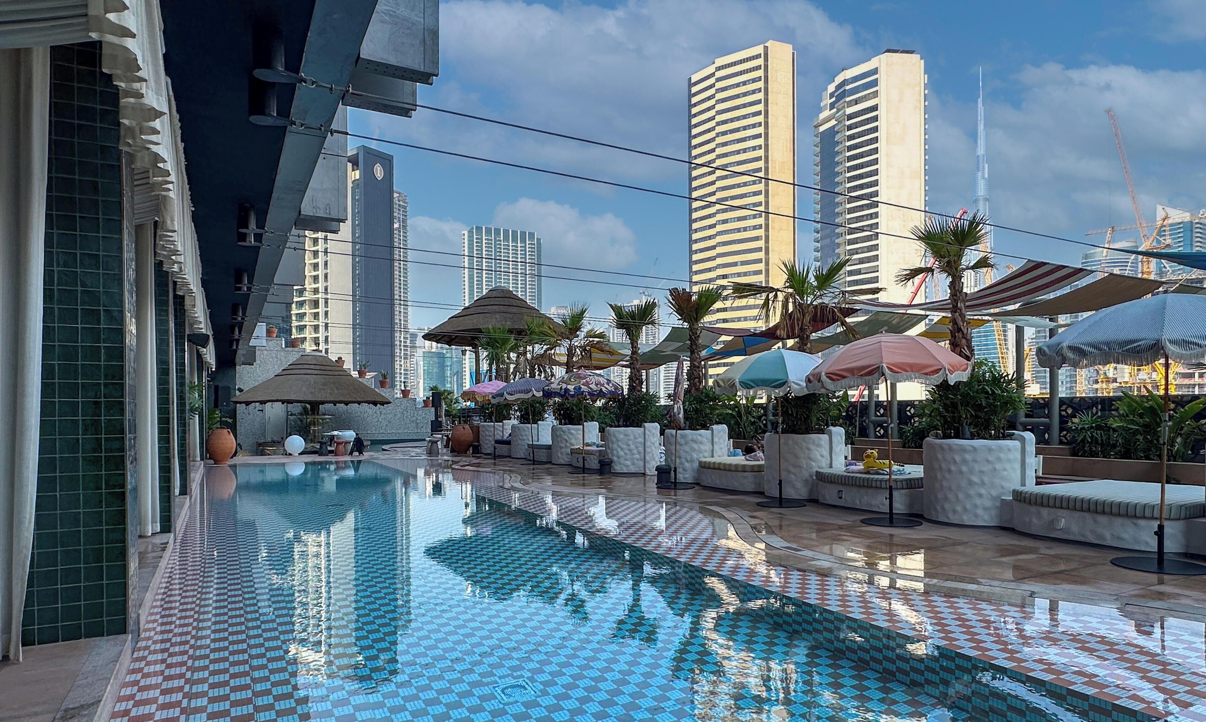 A pool area with tiled flooring, lounge chairs, umbrellas, and tall buildings in the background.
