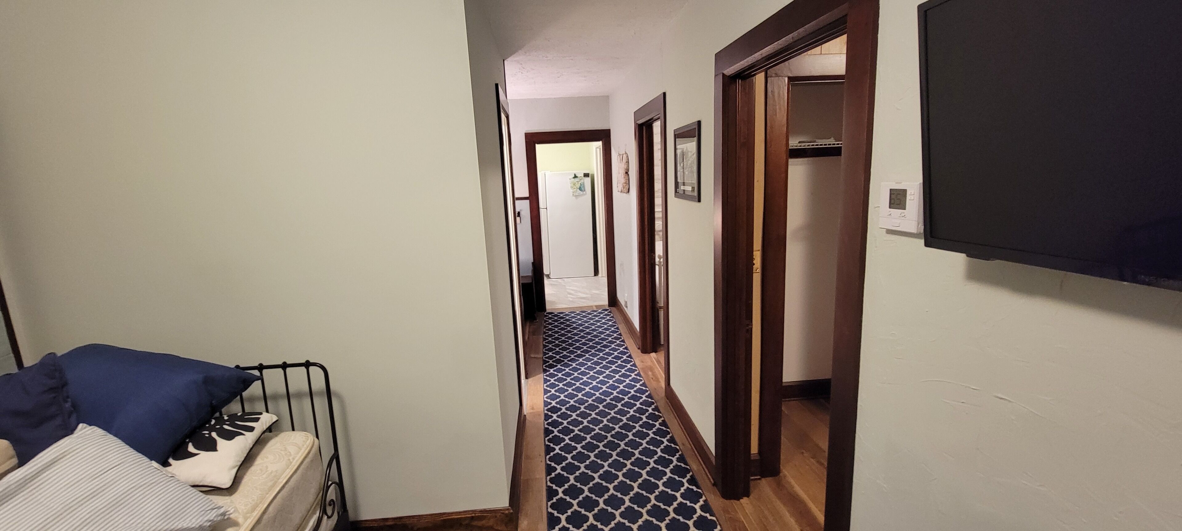 A narrow hallway with a patterned carpet, a bed with pillows, and a television mounted on the wall.