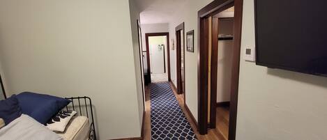 A narrow hallway with a patterned carpet, a bed with pillows, and a television mounted on the wall.