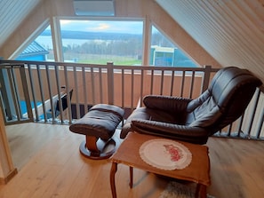 Un balcón con vistas a un lago y montañas, una silla de cuero marrón, una mesita con un mantel individual estampado y un suelo de madera.
