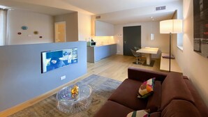 Een moderne woonkamer met een flatscreen-tv, een glazen salontafel, een bruine bank en een keuken in de achtergrond.