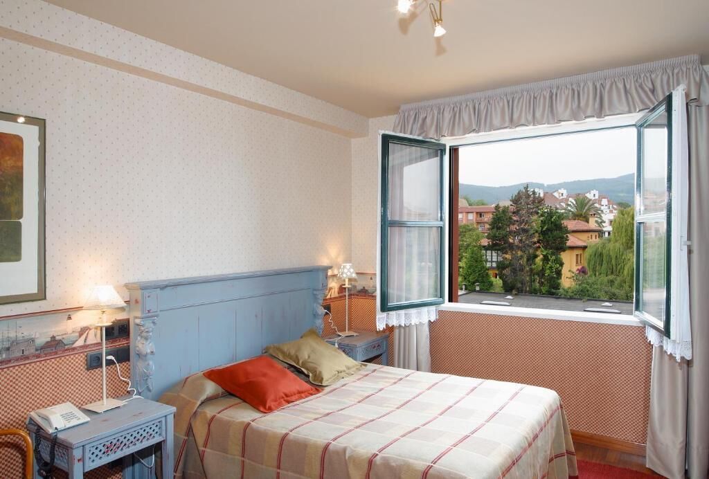 A bedroom with a bed, bedside table, lamp, and a window with an open curtain.