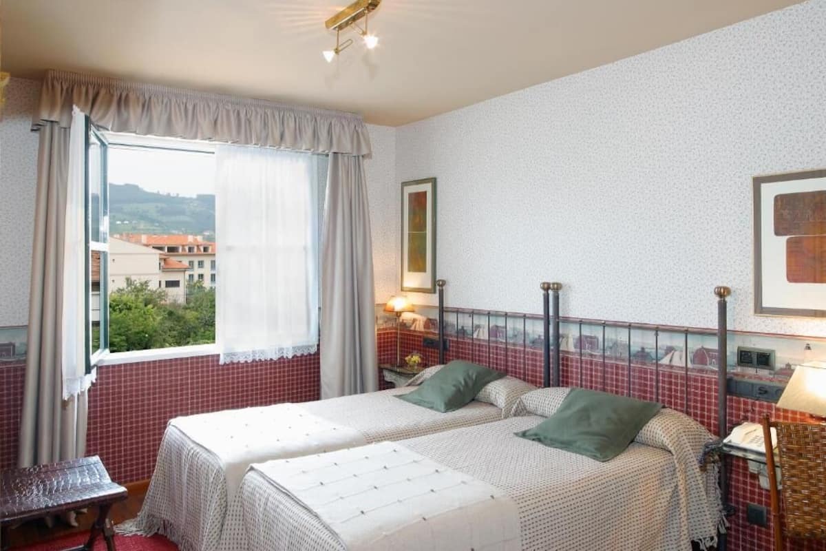 A room with two beds, a window with curtains, a table, and a lamp.