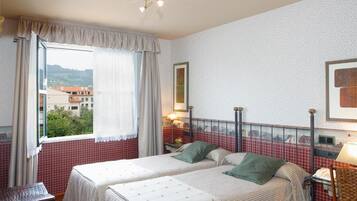 Una habitación con dos camas, una ventana con cortinas, una mesa y una lámpara.