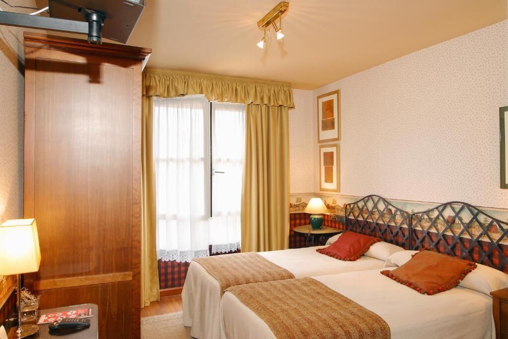 Habitación de hotel con dos camas, un armario de madera, un escritorio con lámpara y una ventana con cortinas.