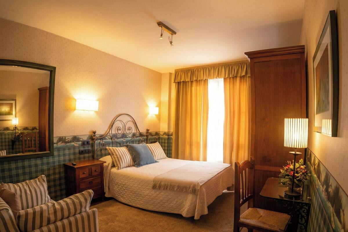 A hotel room with a bed, a chair, a small table, a mirror, and a lamp.