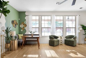 A room with a wooden floor, a dining table with chairs, a sofa, and two armchairs. There are large windows with curtains, a ceiling fan, and wall decorations. - Large Groups' Luxury House Pool Sleep 22 (Round Rock)